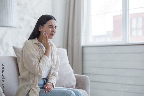 A woman is sick at home alone, has a severe toothache, a brunette is sitting on the sofa by the window, upset