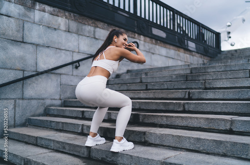 Young woman makes squats to pump the muscles of buttocks and make fit form, simple and effective exercises during morning workout in city. Healthy lifestyle, female keeping the body in sporty shape
