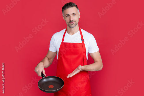 mature man chef in apron with frying pan on red background. presenting product