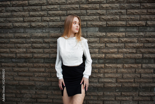 A slender young schoolgirl with blond hair, in a white shirt and a short black skirt, stands against a dark textured stone wall, looks away and keeps her hands on her hips.