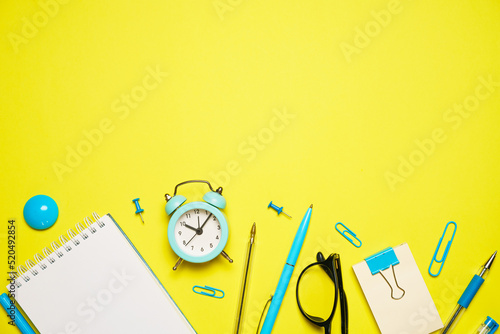 Notebook for writing pen and alarm clock with glasses on yellow background top view, education concept back to school or office supplies