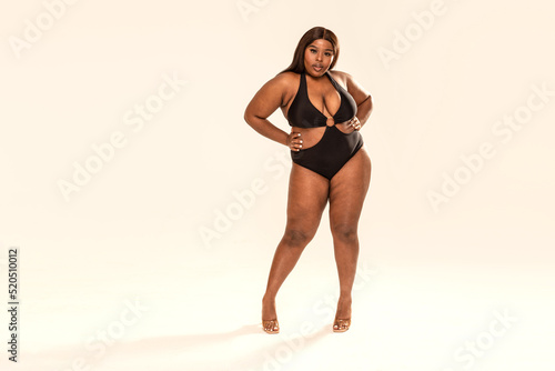 Full-length photo of beautiful plus size dark skinned woman posing on light gray, wearing black fashionable swimsuit.