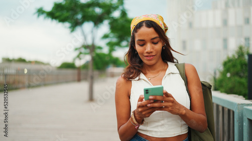 Cute tanned woman with long brown hair wearing white top and yellow bandana walks on bridge with backpack on her shoulder and cell phone in her hand. Girl using phone