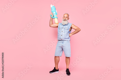 Full length photo of attractive strong man pensioner dressed denim vest rising water gun isolated pink color background