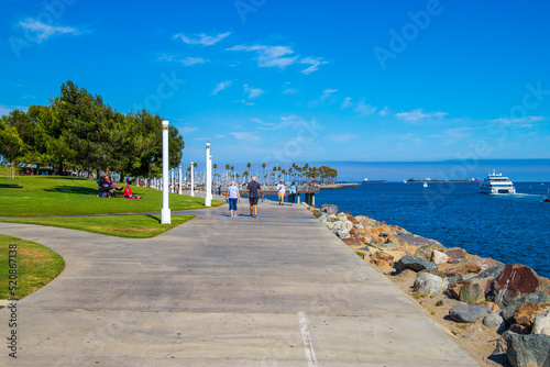 a gorgeous summer landscape in the park with blue ocean water and people relaxing and walking surrounded by lush green trees, grass and plants with blue sky at Shoreline Aquatic Park in Long Beach