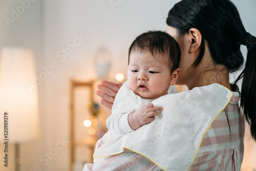 closeup innocent asian infant baby leaning on her mom’s shoulder with cloth is looking away into the space while her mom is burping patting on her back at home.