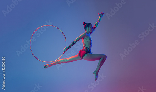 Portrait of young sportive girl, female rhythmic gymnast training, performing with hoop isolated on blue purple background in neon light