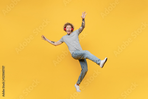 Full body young man 20s he wear grey t-shirt headphones listen to music stand on toes with outstretched hands raise up leg isolated on plain yellow backround studio portrait. People lifestyle concept.
