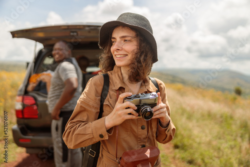Smiling female traveler with photo camera enjoying the journey through the savannah next to safari car