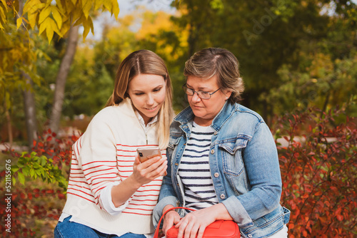 charming women in casual style, mom and daughter are sitting on bench in beautiful autumn park and looking at photos on phone