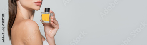 cropped view of woman with bare shoulder holding bottle with perfume isolated on grey, banner