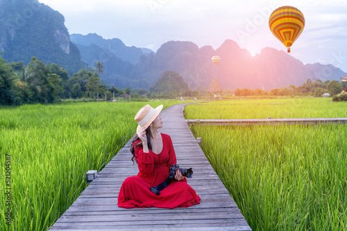 A woman holding a camera sits on a wooden walkway and looks at balloons with green rice fields in Vang Vieng, Laos.