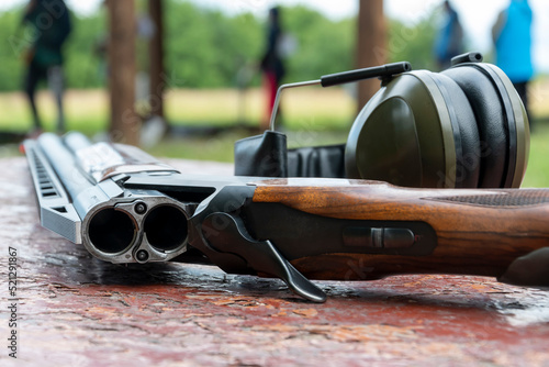 A sports double-barreled shotgun and headphones for shooting lie on a wooden table against the background of the shooting range.