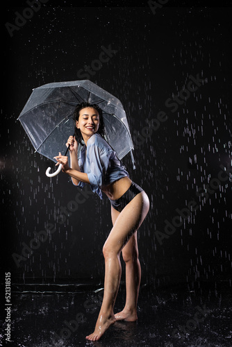 Beautiful girl in a black swimsuit and shirt. Stands in the rain, holds a transparent umbrella in his hands. Posing playfully. Water splash on black background. Aqua studio