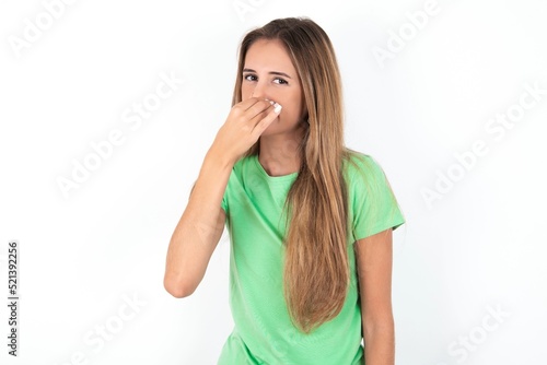 young beautiful woman wearing green T-shirt over white background, smelling something stinky and disgusting, intolerable smell, holding breath with fingers on nose. Bad smell