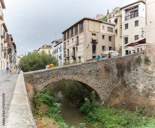 View at the Darro Street, Paseo de los tristes, cabrera bridge, touristic people visiting iconic and picturesque street, Alhambra fortress as background, in Granada, Spain