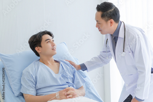 Senior doctor and young male patient who lie on the bed while checking pulse, consult and explain with nurse taking note and supporting in hospital wards.