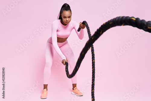 Confident young African woman exercising with battle ropes against pink background