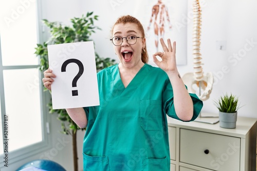 Young redhead physiotherapist woman holding question mark at the clinic doing ok sign with fingers, smiling friendly gesturing excellent symbol