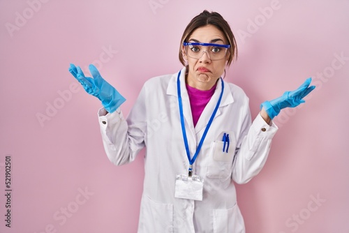 Hispanic woman wearing scientist uniform clueless and confused expression with arms and hands raised. doubt concept.