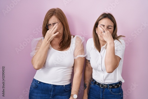 Hispanic mother and daughter wearing casual white t shirt over pink background smelling something stinky and disgusting, intolerable smell, holding breath with fingers on nose. bad smell