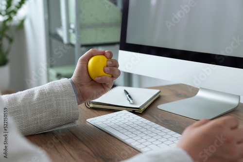 Man squeezing antistress ball while working with computer in office, closeup