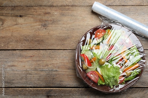 Bowl of fresh salad with plastic food wrap on wooden table, flat lay. Space for text