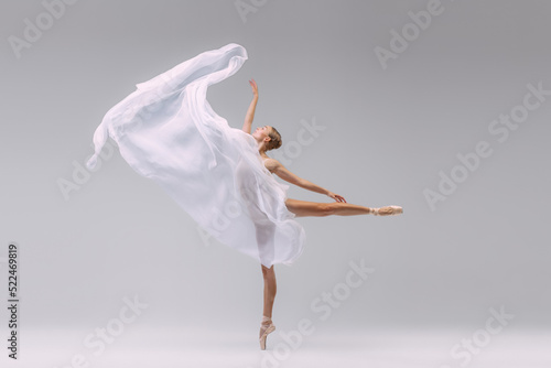 Portrait of young ballerina dancing with fabric isolated over grey studio background. Standing on tiptoe and throwing transparent cloth