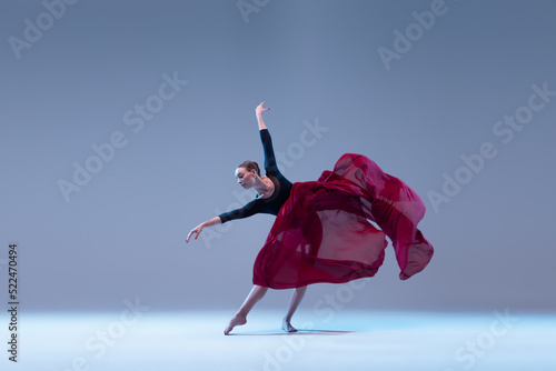 Portrait of young ballerina dancing with deep red fabric isolated over blue grey studio background. Artistic performance of classic dance