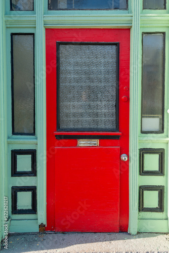 The exterior facade of a vintage shop with an orange wooden door with a glass window, mail slot and door handle. The door is on a lime green building with black trim. here's a transom window over door