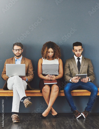 Group of design interns on wireless online devices waiting at a job interview of a startup company. Diverse people sitting, waiting for a meeting with human resources for a job
