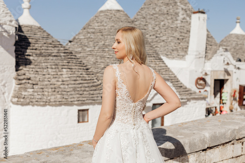 Back view of young happy woman bride standing near conical roofs of trullo in sunny Italy, putting hand on waist posing.