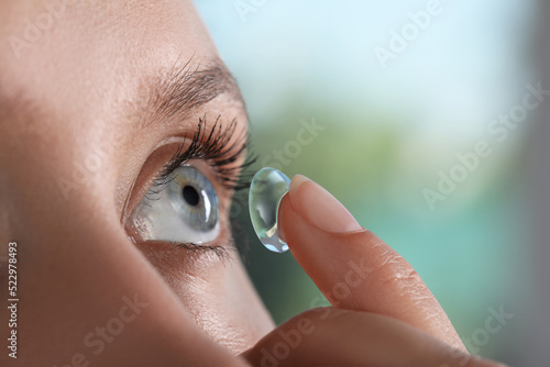 Young woman putting contact lens in her eye on blurred background, closeup