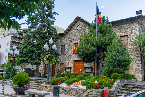 Sant Julia de Loria Town Hall in Andorra, a stone building with the Andorran flag on a mast and a garden with trees and flowers in summer, with the mountains behind and closed wooden doors