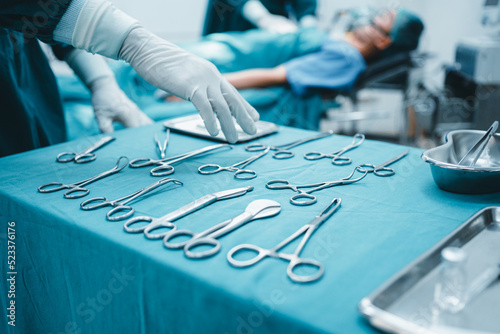 Close up surgeon doctor's hand with hygiene glove taking sterile surgical instrument tool, equipment in operating room patient in background for surgery. Hospital, medical healthcare emergency concept