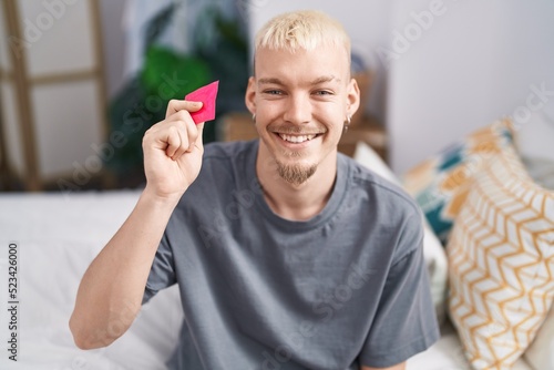 Young caucasian man holding condom sitting on bed looking positive and happy standing and smiling with a confident smile showing teeth