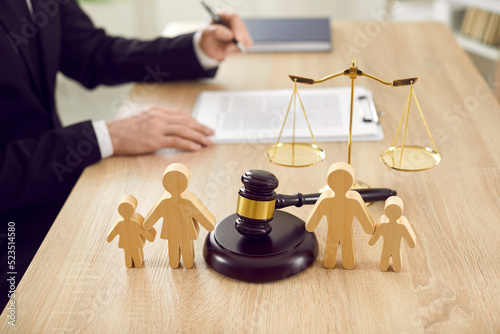 Figures of family are separated by judge's gavel, which symbolizes divorce case and custody of child. Close up of wooden figurines of people on background of judges and scales of justice.