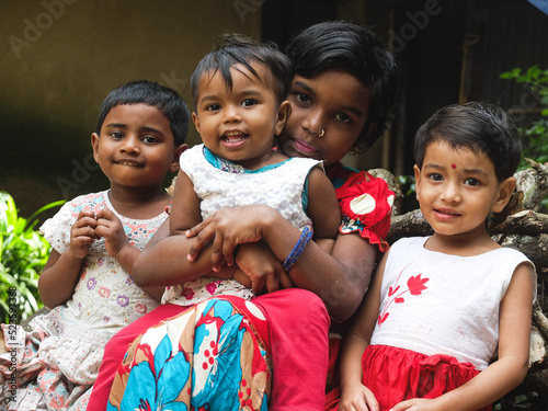 South asian sisters sitting in front of the door of their mud made home at village, Bangladeshi kids playing outside