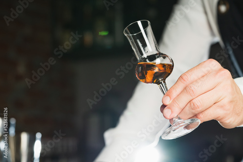 Strong alcohol. A professional bartender in a white shirt and black apron holds a tasting glass, the trend drinks concept.