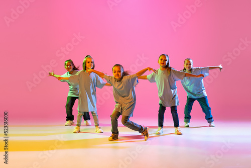 Group of children, little girls in sportive casual style clothes dancing in choreography class isolated on orange background in purple neon light.