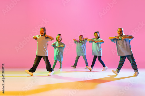 Group of children, little girls in sportive casual style clothes dancing in choreography class isolated on orange background in purple neon light.