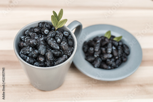 Kamchatka berries / haskap berries / honeysuckle (Lonicera caerulea) in a gray cup and on a gray plate decorated with leaves, placed on a wooden board, side view.