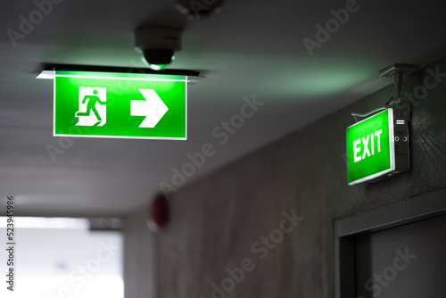 A green fire exit sign is placed on the ceiling along the dimly lit corridor and there is green exit sign on the exit door.