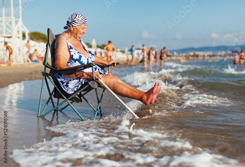 Old white woman in a swimsuit and a bandana sits in a tourist chair near the Black Sea near the city of Anaps and meets the oncoming wave