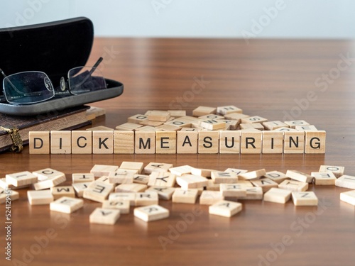 dick measuring word or concept represented by wooden letter tiles on a wooden table with glasses and a book