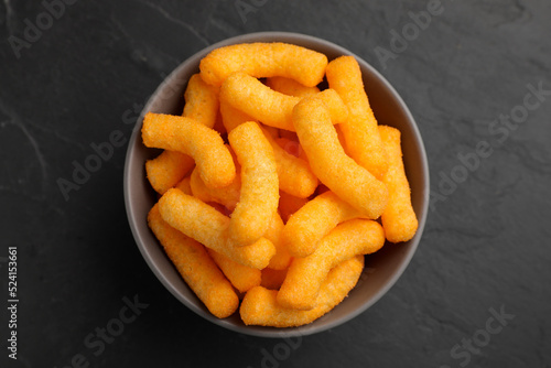 Many tasty cheesy corn puffs in bowl on black table, top view