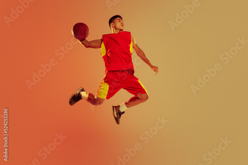 Portrait of muscular young man, basketball player in motion, throwing ball in a jump isolated over orange studio background in neon light
