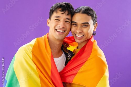 Portrait of happy biracial male couple embracing and holding lgbt flag on purple background