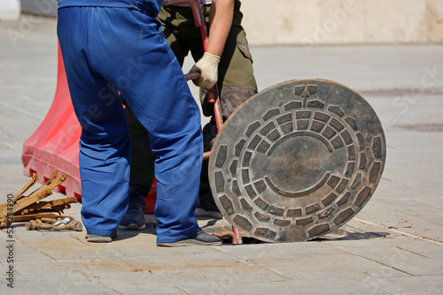 Two workers standing over the open sewer hatch on a street and holding metal ladder. Concept of repair of sewage, underground utilities, water supply system, cable laying, water pipe accident