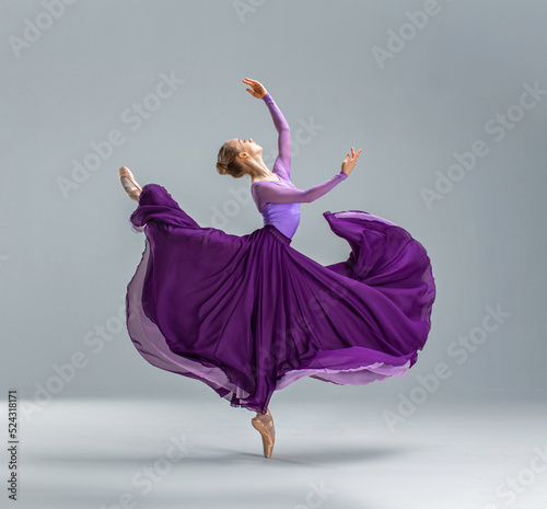 Ballerina in purple ballet leotard and violet long ballet skirt dancing in white studio on pointe shoes.